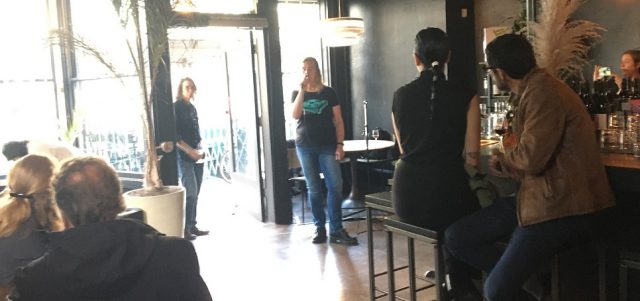 Interior of a bar. Several people listen to a woman who is speaking into a microphone.