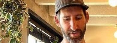 a man faces the camera and looks down. he is wearing a black t-shirt an d a baseball style hat.