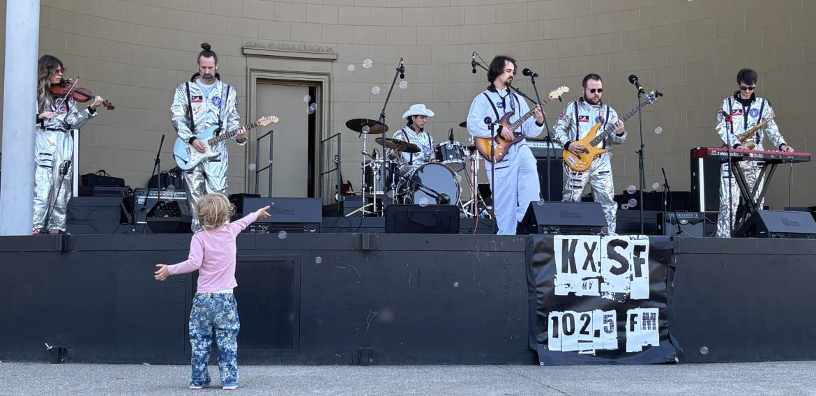 a rock band performs on a stage while dressed as astronauts. in the foreground a young girl gestures out to them